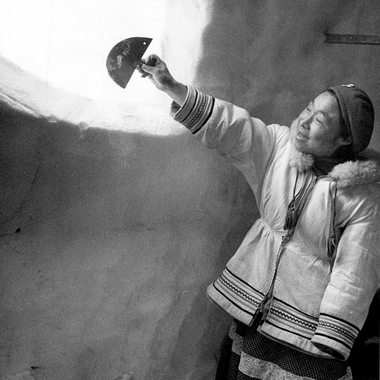 Photo en noir et blanc d'une femme en parka et chapeau tenant un couteau en forme de demi-cercle et le pointant vers une fenêtre ouverte découpée dans un igloo.