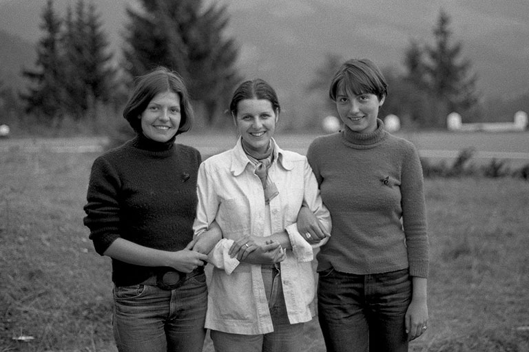 Photo noir et blanc de trois jeunes femmes se tenant par les coudes dans un paysage montagneux.