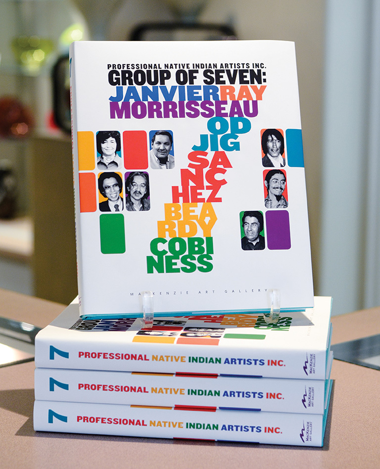 One book sits on a stand on top of three books in a pile on their sides. The book title is 7: Professional Native Indian Artists Inc.