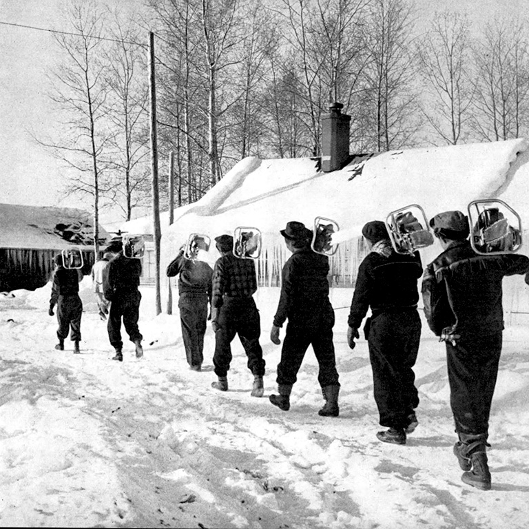 Photo en noir et blanc de sept hommes marchant dans la neige avec des tronçonneuses sur les épaules.