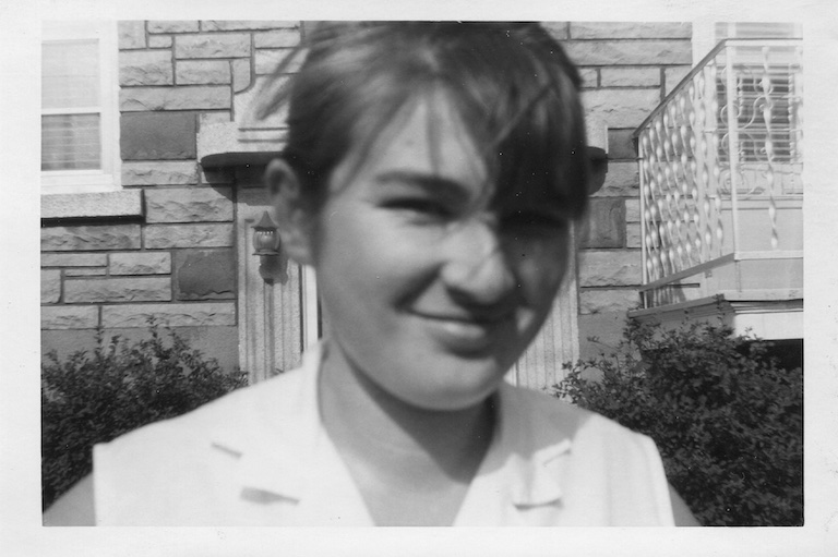 Photo en noir et blanc en gros plan d’une jeune fille souriante. Derrière elle, un immeuble à appartements.