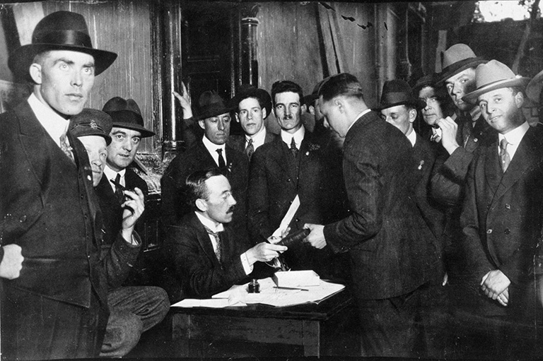 A group of men in black suits standing around a man sitting at a table with papers and a pen.