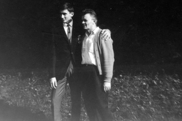 Photo en noir et blanc d’un jeune homme tenant son père par les épaules. Ils sont à l’extérieur, des arbres derrière eux.