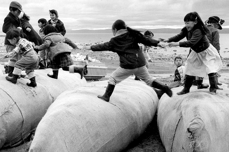 Une photo en noir et blanc d'enfants en manteaux et bottes courant sur de grands sacs de fournitures.