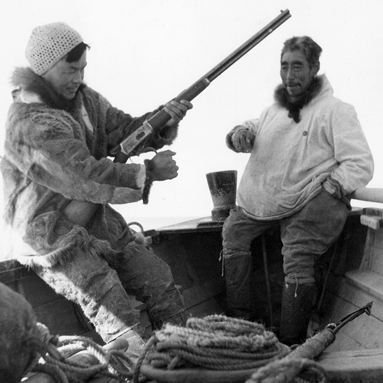 Photo en noir et blanc de deux hommes en parkas assis sur un bateau. L'un des hommes tient un fusil.