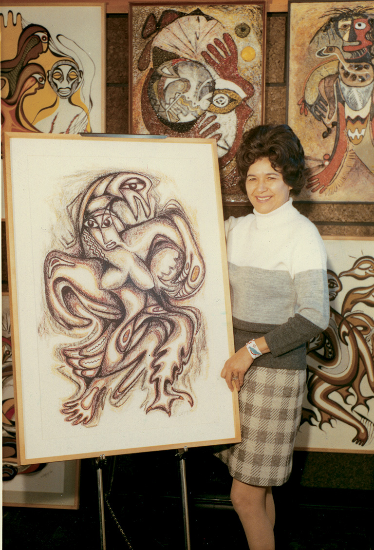 A woman in a skirt and sweater stands next to a painting showing a contorted body painted in an abstract manner. There are paintings on the wall behind the woman.