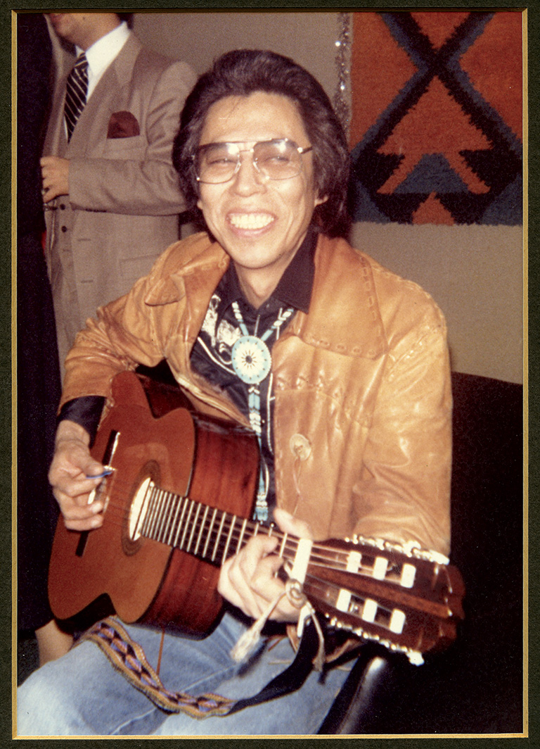 A man in a leather jacket and large glasses sits and strums a guitar.