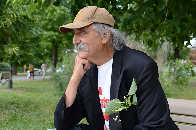 Photo couleur d’un homme de profil en plan taille. Il est assis sur un banc dans un parc, il porte une casquette.