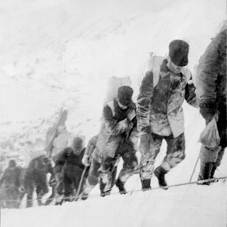 Black and white photo of six men in climbing gear with backpacks hiking up a snowy trail.