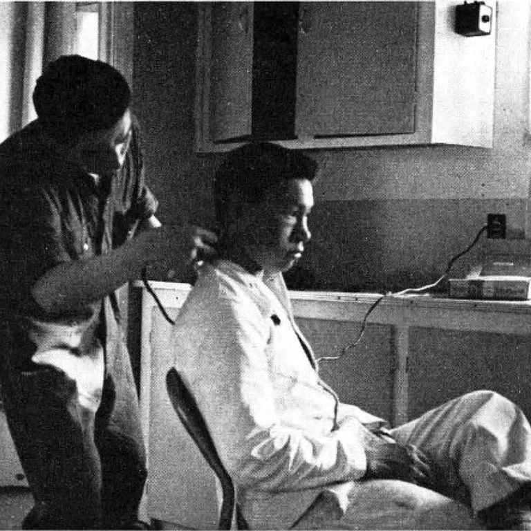 Une photo d'un homme assis et d'un autre homme se tenant derrière et utilisant un rasoir à cheveux.