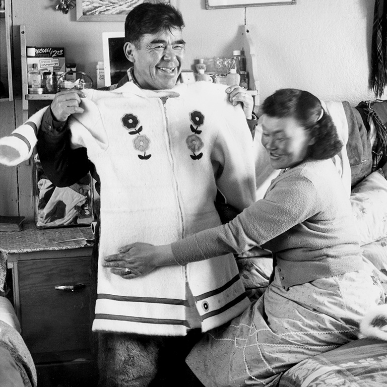 Un homme tient une parka blanche en travers de sa poitrine pour montrer la taille. Une femme assise à côté de l'homme l'aide.